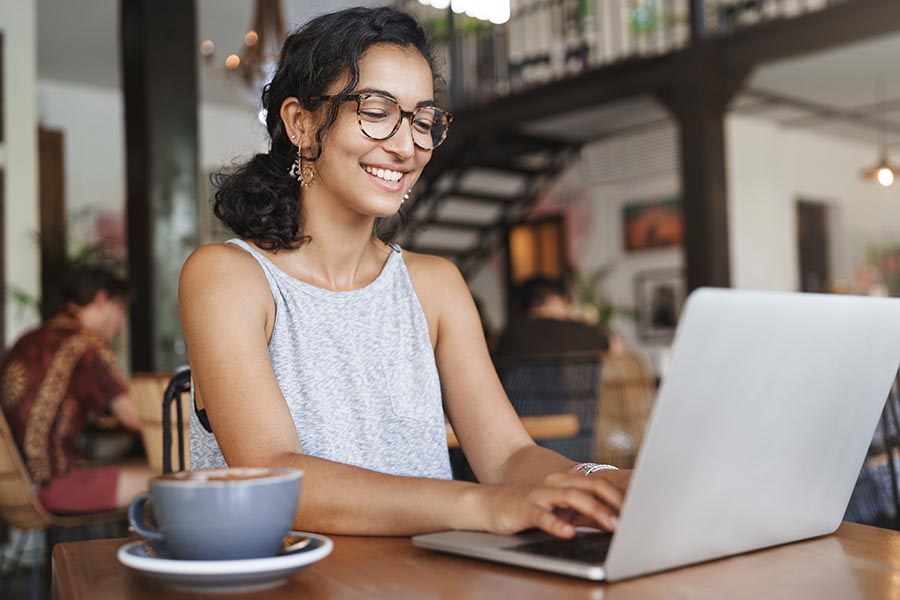 Client Center - Young Woman in a Bustling Cafe Smiles and Uses Her Laptop With a Cappuccino Beside Her