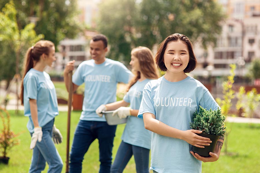 Specialized Business Insurance - Young Woman Holding a Potted Shrub and Wearing a Volunteer Shirt Smiles While Fellow Volunteers Chat Behind Her